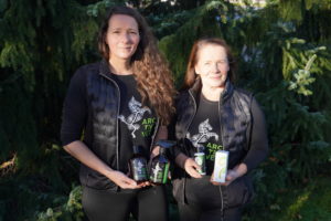 Kaksi naista seisoo kuusien edessä ulkona ja pitelee yrityksensä tuotteita käsissään.